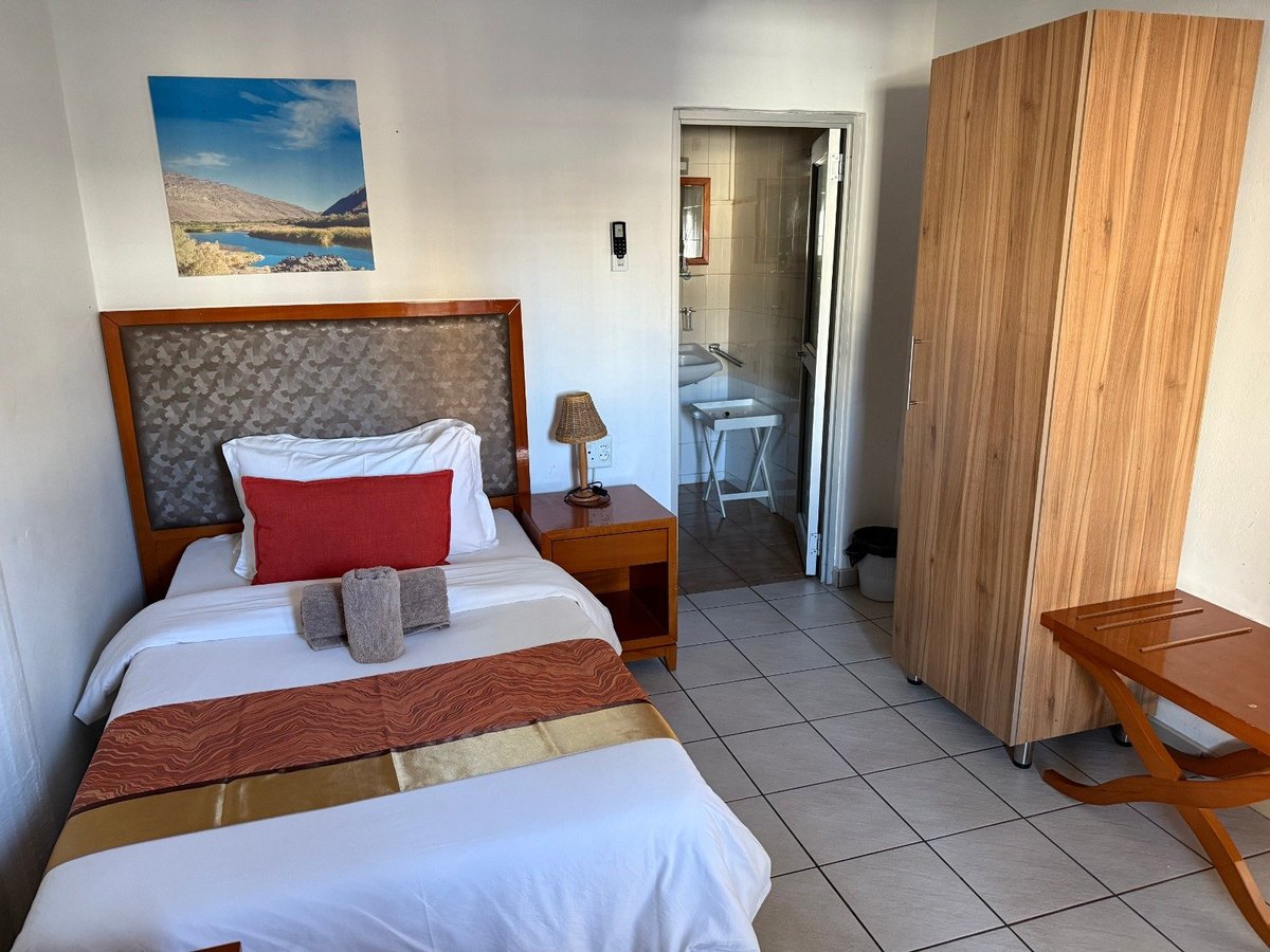 Hotel bedroom with single bed, red pillow, wooden headboard, bedside table, coastal photo on wall, wooden wardrobe, and doorway to bathroom
