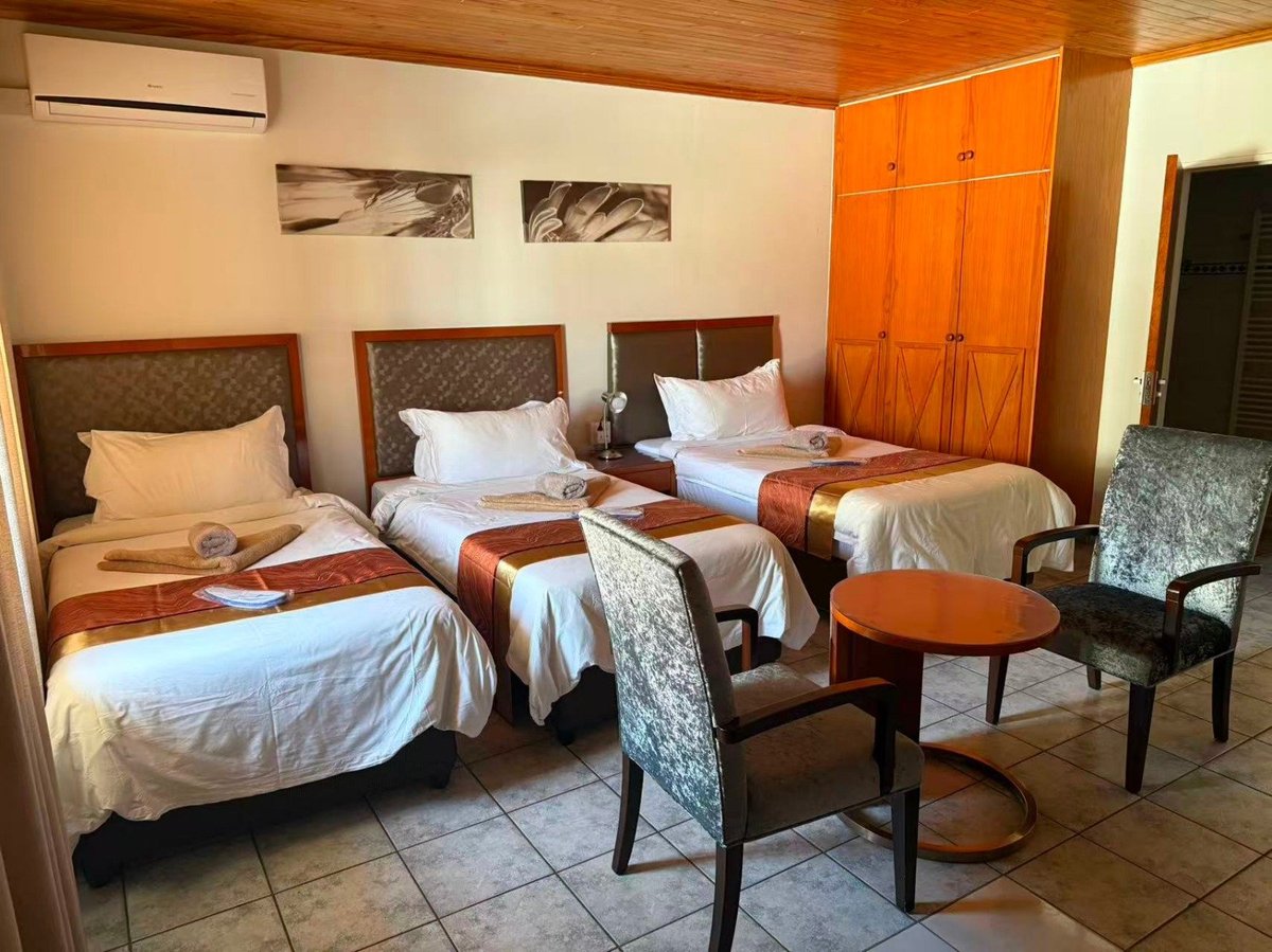 Hotel bedroom with three single beds, wooden furniture, orange wardrobe, and seating area with black chairs.